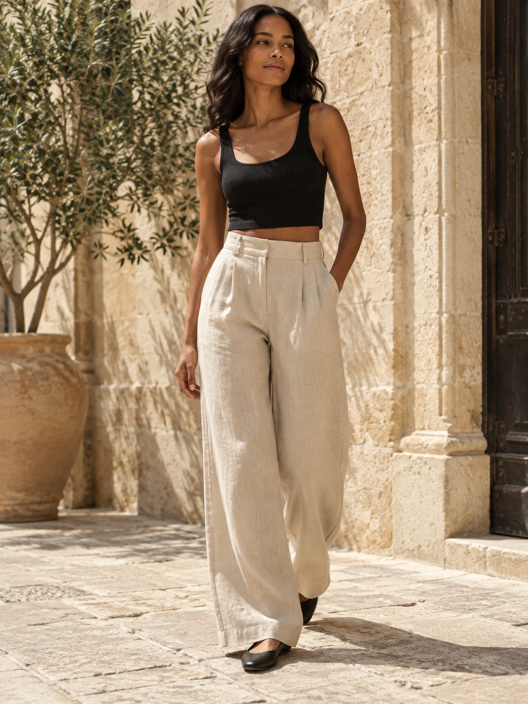 Adult model wearing relaxed sand-coloured linen trousers in a sunlit courtyard